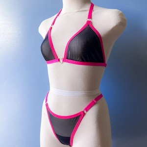 Puede incluir: Conjunto de lencería negro con top halter y tanga. El conjunto presenta una tela negra transparente con ribetes y detalles en rosa brillante. El top tiene un detalle en forma de corazón en el centro. El tanga tiene un ribete rosa a juego.