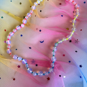 May include: A beaded necklace featuring iridescent, round beads with colorful accents. The necklace has a silver clasp and is strung with neon pink, yellow, and blue thread. The beads are spaced with small, brightly colored beads. The necklace is displayed on a colorful, star-speckled background.