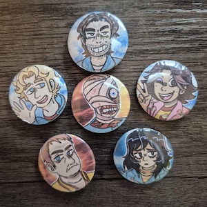 Könnte beinhalten: Eine Sammlung von sieben runden, farbenfrohen Ansteckbuttons mit Cartoon-Porträts. Die Illustrationen zeigen verschiedene Charaktere mit unterschiedlichen Gesichtsausdrücken und Stilen, vor blau-orangen Hintergründen. Die Buttons sind auf einer Holzoberfläche angeordnet.