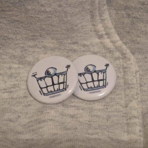 Puede incluir: Dos botones redondos blancos con un diseño de carrito de la compra de dibujos animados. El carrito tiene una bola azul y una sonrisa con dientes. Los botones están sobre un fondo de tela gris.