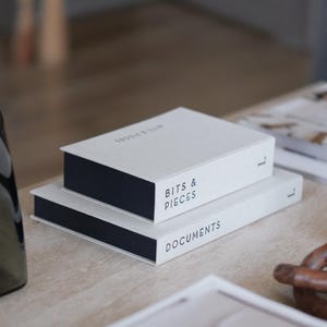 Op de afbeelding: Twee witte boeken met donkerblauwe ruggen, met de tekst "BITS & PIECES" en "DOCUMENTS", liggen op een lichtgekleurd houten oppervlak. Andere boeken en een decoratief object zijn ook zichtbaar.