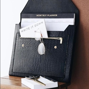 Könnte beinhalten: Schwarze Clutch aus Kunstleder in Briefumschlagform mit einem goldenen Reißverschluss und einem goldenen Druckknopfverschluss. Die Clutch hat ein Kroko-Prägemuster und ist geöffnet, um einen weißen Monatskalender mit dem Text "MONTHLY PLANNER" und einen weißen Notizblock mit dem Text "WRITE IT DOWN" zu zeigen.