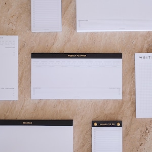Könnte beinhalten: Eine Auswahl weißer Notizblöcke mit schwarzen Rändern und goldfarbener Schrift. Die Blöcke tragen die Aufschriften "Things to Do", "Deskpad" und "Weekly Planner". Sie liegen auf einer hellbraunen Oberfläche.