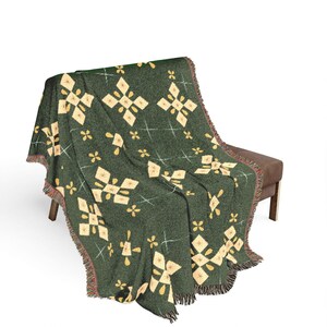 May include: A dark green woven throw blanket featuring a repeating pattern of cream-colored diamond shapes and small four-petaled floral motifs, accented by thin white cross-stitch details and finished with a fringed edge along the sides.