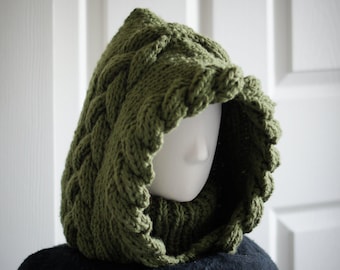 May include: An olive green knitted hood with a thick cable knit pattern along the edges and a ribbed neck section. The hood is displayed on a white mannequin head, showcasing the textured yarn construction and the deep, rounded shape of the piece.