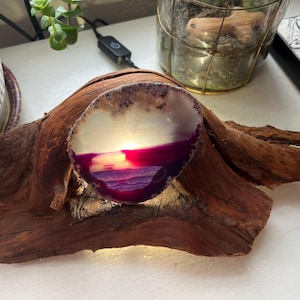 May include: A purple and white agate geode with a sunset scene inside is lit from behind and mounted on a piece of driftwood.