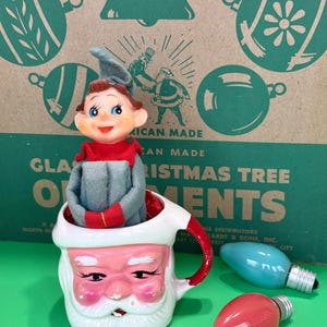 May include: A vintage Christmas decoration featuring a ceramic Santa mug holding an elf figurine. The mug has a pink face and white beard. Two colored light bulbs are next to the mug. The background has a green Christmas tree ornament design.