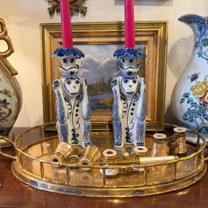 May include: Two blue and white monkey-shaped candle holders with pink candles. The candle holders are on a gold-colored tray with small gold-colored binoculars and other decorative items. A decorative vase is on the right.