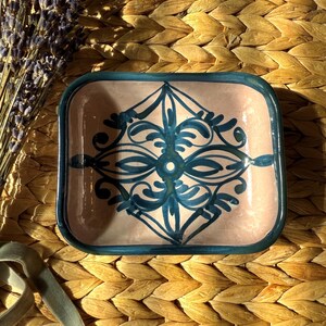 May include: A rectangular ceramic dish with a pink interior and a teal blue border. The dish features a symmetrical teal blue floral design. The dish is displayed on a woven straw mat, with a sprig of lavender and a green ribbon nearby.