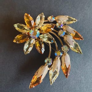 May include: Vintage floral brooch with a gold-tone metal base. The brooch features a large flower with amber-colored crystal petals and iridescent center stones. Additional amber and green crystal leaves and accents complete the design.
