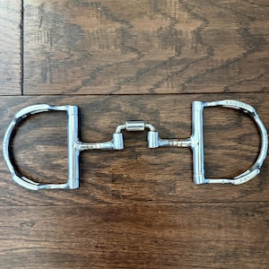 May include: A silver-colored metal horse bit featuring D-shaped cheekpieces and a central jointed mouthpiece with copper-colored accents. The right cheekpiece is engraved with the number 620239381 and the text PAT.