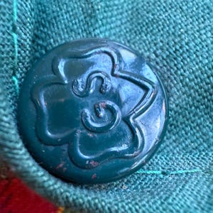 May include: Close-up of a dark teal button with a floral design, attached to a green fabric. The button is round and has a raised, stylized flower pattern. The fabric is a textured weave, with a red and yellow plaid pattern visible.