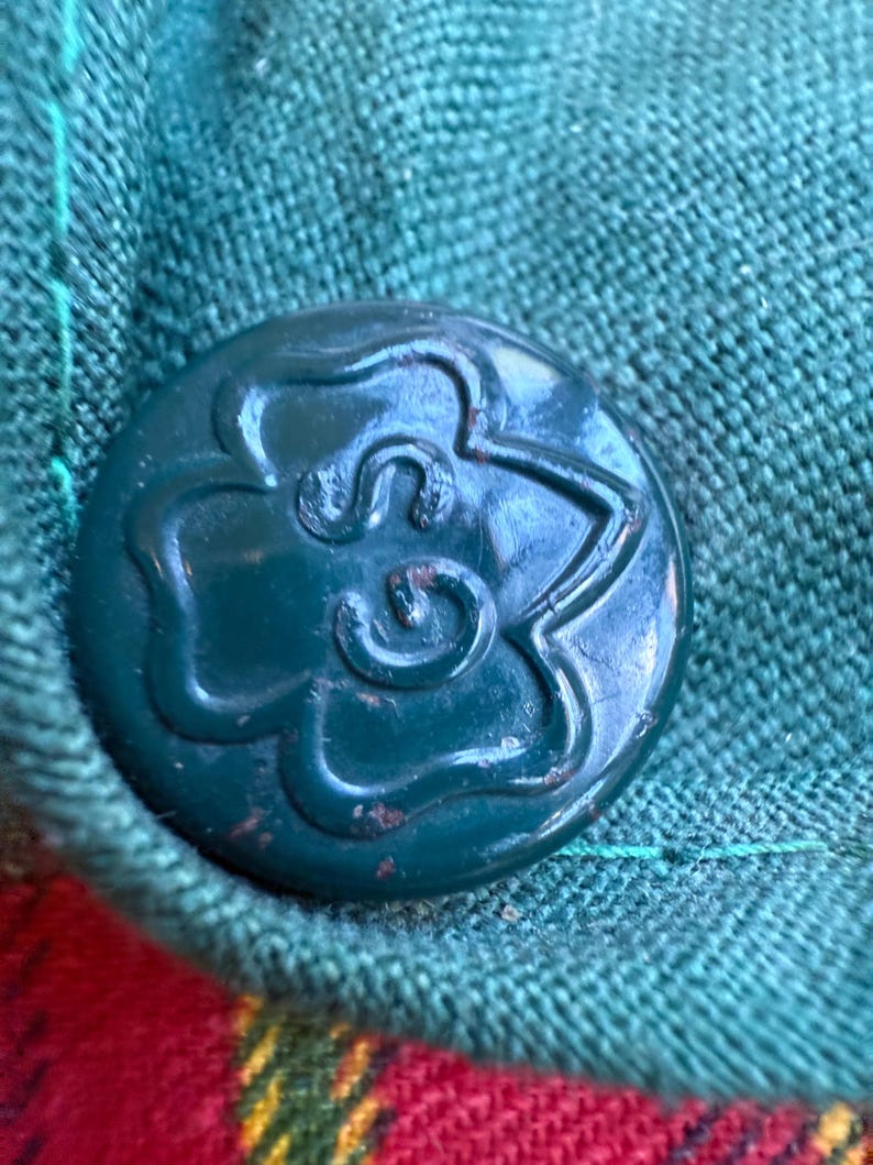May include: Close-up of a dark teal button with a floral design, attached to a green fabric. The button is round and has a raised, stylized flower pattern. The fabric is a textured weave, with a red and yellow plaid pattern visible.