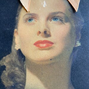 May include: Vintage portrait of a woman with blue eyes and red lipstick. The image has a soft focus and is set against a dark background. A peach-colored ribbon is attached to the top of the image.
