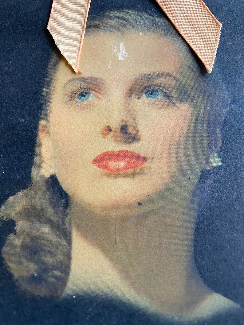 May include: Vintage portrait of a woman with blue eyes and red lipstick. The image has a soft focus and is set against a dark background. A peach-colored ribbon is attached to the top of the image.