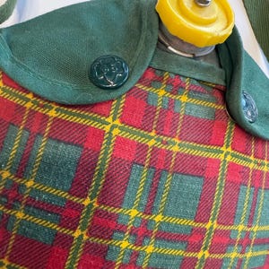 May include: A vintage canteen with a red, green, and yellow plaid pattern. The canteen has a green flap with a green button and a yellow cap. The canteen has a green strap.