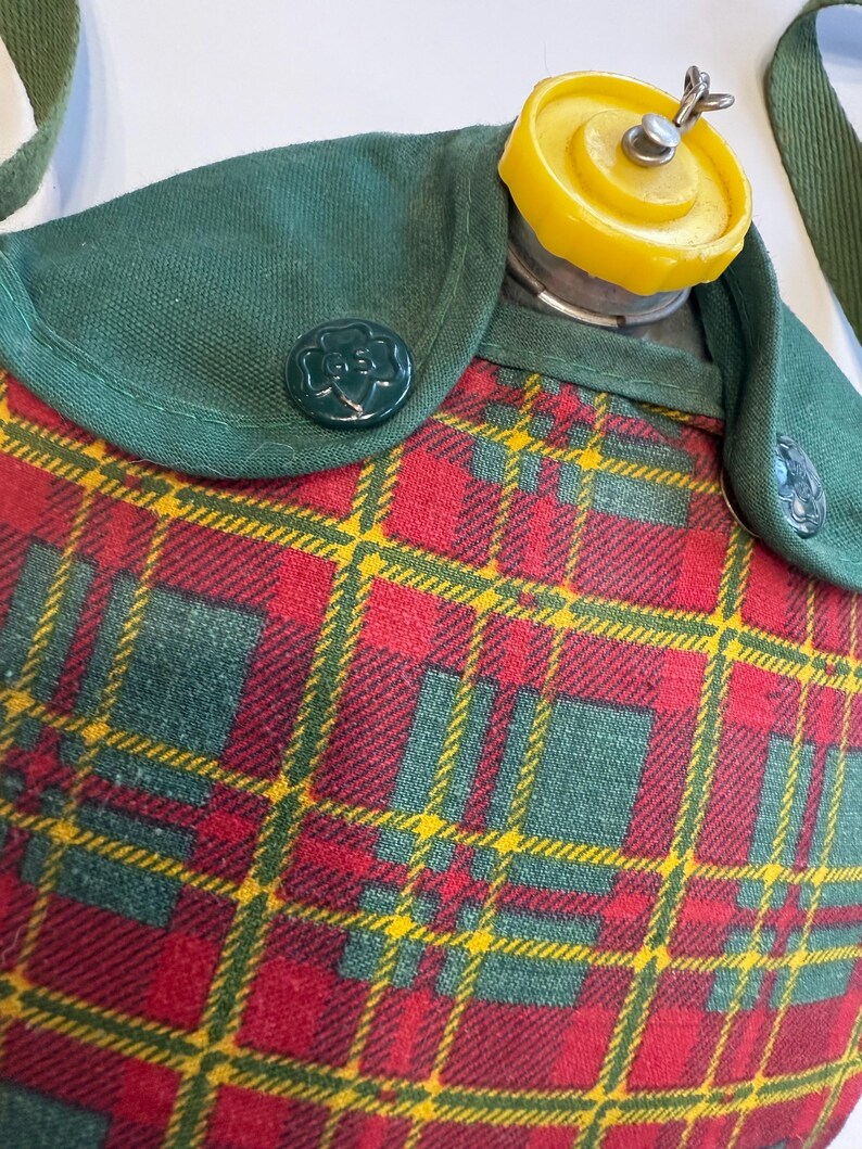 May include: A vintage canteen with a red, green, and yellow plaid pattern. The canteen has a green flap with a green button and a yellow cap. The canteen has a green strap.