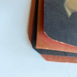 May include: A stack of vintage photographs with a black and red border. The edges of the photos are slightly worn, and the colors are faded. The top photo has a dark background with a light-colored area.