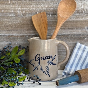 May include: A tan ceramic pitcher with a handle, featuring the word "Grease" in blue script and a decorative design. Two wooden utensils are inside the pitcher. A blue and white striped cloth and a rolling pin are also visible.
