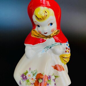 May include: A vintage ceramic figurine of a girl in a red hooded dress and white skirt. She is holding a basket of flowers and has a floral pattern on her skirt.