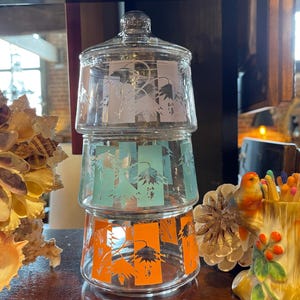May include: A set of three glass canisters with lids. The canisters are stacked on top of each other. The bottom canister has an orange floral design, the middle canister has a blue floral design, and the top canister has a white floral design.