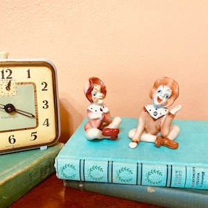 May include: Two small ceramic figurines of clowns, one wearing a pink and white outfit and the other wearing a blue and white outfit. The figurines are sitting on top of a stack of books.