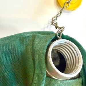 May include: Close-up of a green fabric bag with a metal spring and a yellow cap attached by a chain. The bag has a plaid pattern in red, yellow, and green. A green button is visible on the bag.