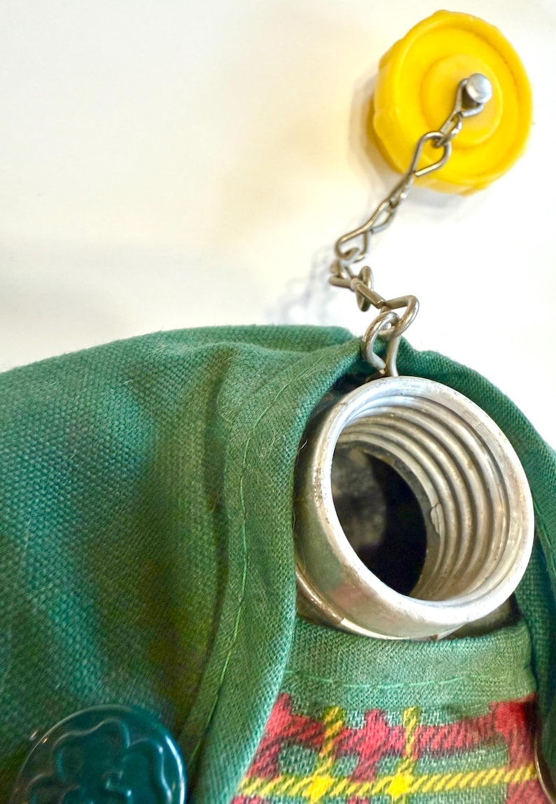 May include: Close-up of a green fabric bag with a metal spring and a yellow cap attached by a chain. The bag has a plaid pattern in red, yellow, and green. A green button is visible on the bag.