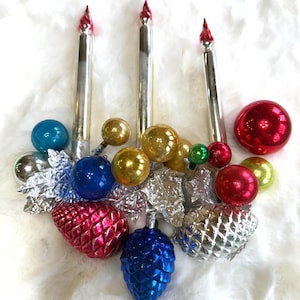May include: A collection of vintage Christmas ornaments and candle decorations. The arrangement includes silver candle-shaped ornaments with red tips, along with various colorful glass ball ornaments and pinecone-shaped ornaments in red, blue, and silver.