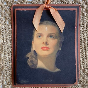 May include: Vintage print of a woman with blue eyes and red lips, titled "Stardust." The image is set against a black background and framed with a brown border. A peach-colored ribbon is tied at the top. Copyright information is visible at the bottom.