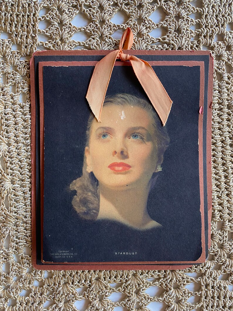 May include: Vintage print of a woman with blue eyes and red lips, titled "Stardust." The image is set against a black background and framed with a brown border. A peach-colored ribbon is tied at the top. Copyright information is visible at the bottom.