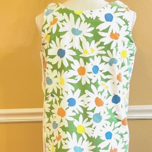 May include: A sleeveless top featuring a vibrant green background covered in a repeating pattern of white daisy flowers. The centers of the daisies are filled with solid circles in shades of blue, yellow, and orange, creating a colorful floral design.