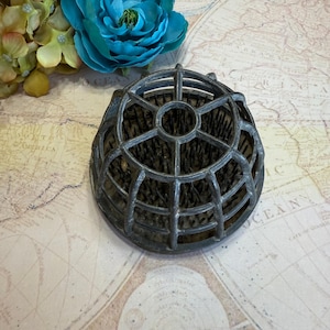 May include: A vintage metal flower frog with a cage-like design, featuring numerous pin-like holders for arranging flowers. The frog is a dull silver color and sits on a map-themed background, with a blue flower in the upper left corner.