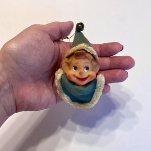 May include: A vintage Christmas ornament featuring a smiling elf with a blue and white hat and a bell on top. The elf has a brown face, blue eyes, and a red mouth.