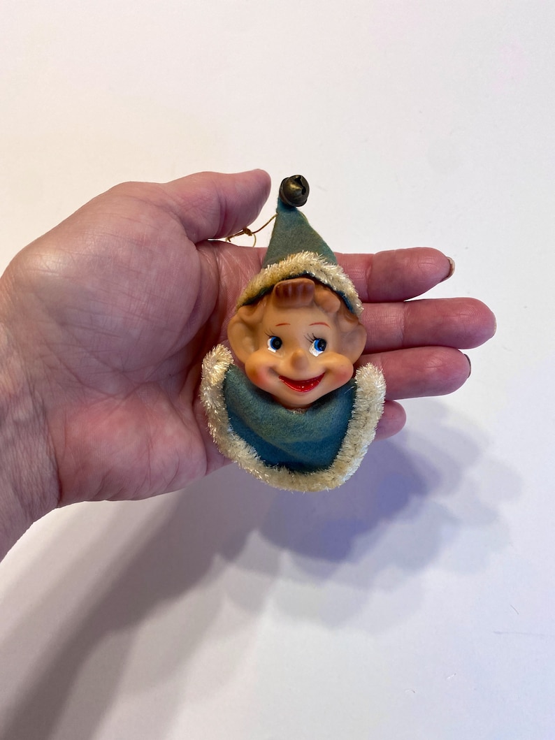 May include: A vintage Christmas ornament featuring a smiling elf with a blue and white hat and a bell on top. The elf has a brown face, blue eyes, and a red mouth.