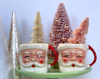 May include: Two white ceramic Santa Claus mugs with red trim and a white beard. The mugs are sitting on a green pedestal cake stand.