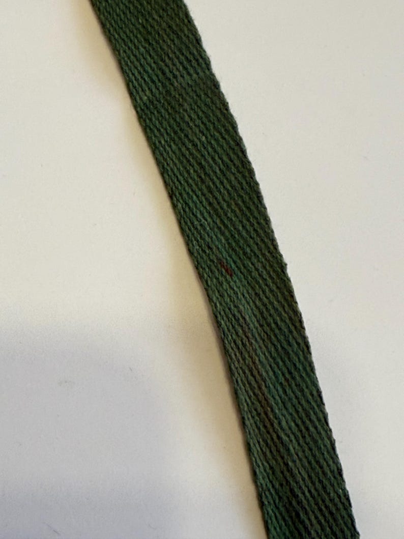 May include: Close-up of a dark green woven strap. The strap has a diagonal pattern and appears to be made of a textured material. A few small, dark red marks are visible on the strap.