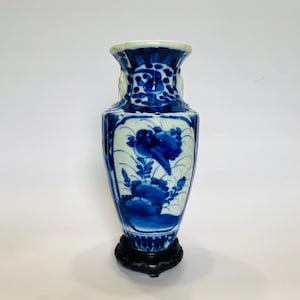 May include: A blue and white porcelain vase with a floral design and a bird. The vase is on a wooden stand.