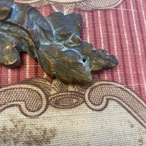 K&ouml;nnte beinhalten: Ein bronzefarbenes, blattf&ouml;rmiges Dekorationsst&uuml;ck mit einem Loch in der Mitte. Das St&uuml;ck befindet sich auf einem gemusterten Stoffhintergrund.