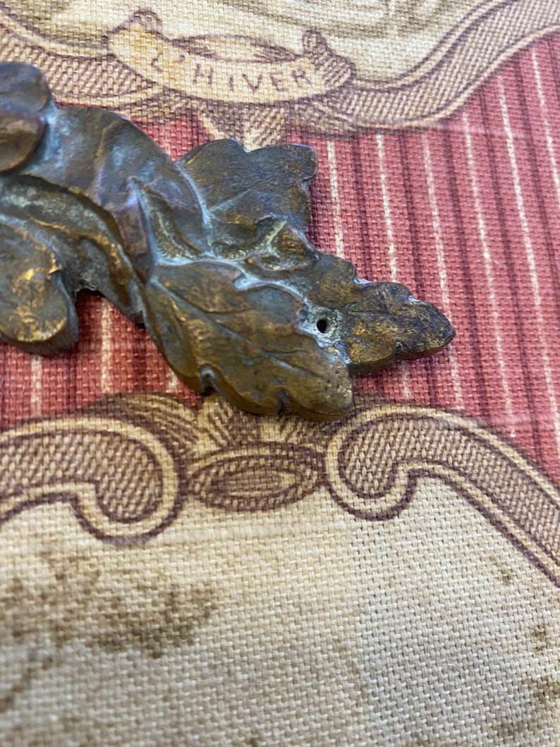 K&ouml;nnte beinhalten: Ein bronzefarbenes, blattf&ouml;rmiges Dekorationsst&uuml;ck mit einem Loch in der Mitte. Das St&uuml;ck befindet sich auf einem gemusterten Stoffhintergrund.