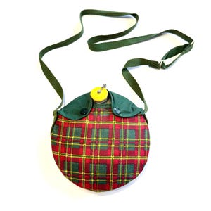 May include: A vintage, round canteen with a red, green, and yellow plaid pattern. The canteen has a green fabric top with a yellow cap and a green strap for carrying. The canteen is designed to be worn over the shoulder.