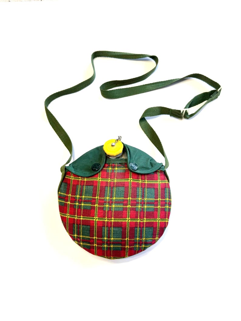 May include: A vintage, round canteen with a red, green, and yellow plaid pattern. The canteen has a green fabric top with a yellow cap and a green strap for carrying. The canteen is designed to be worn over the shoulder.