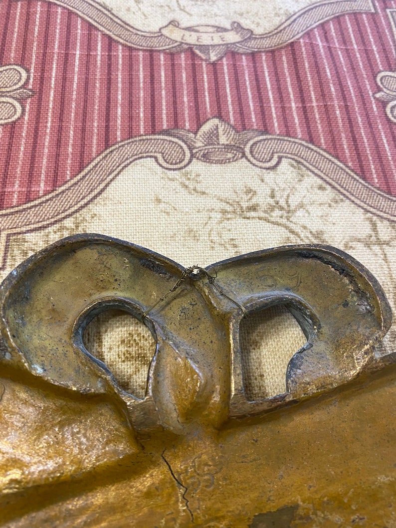 K&ouml;nnte beinhalten: Ein goldfarbener Metallrahmen mit einem dekorativen Design. Der Rahmen hat zwei &Ouml;ffnungen auf der R&uuml;ckseite, wahrscheinlich zum Aufh&auml;ngen.