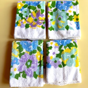 May include: Four white hand towels with a floral pattern in shades of blue, purple, yellow, and green. The towels have a white border at the bottom. The towels are arranged on a yellow background.