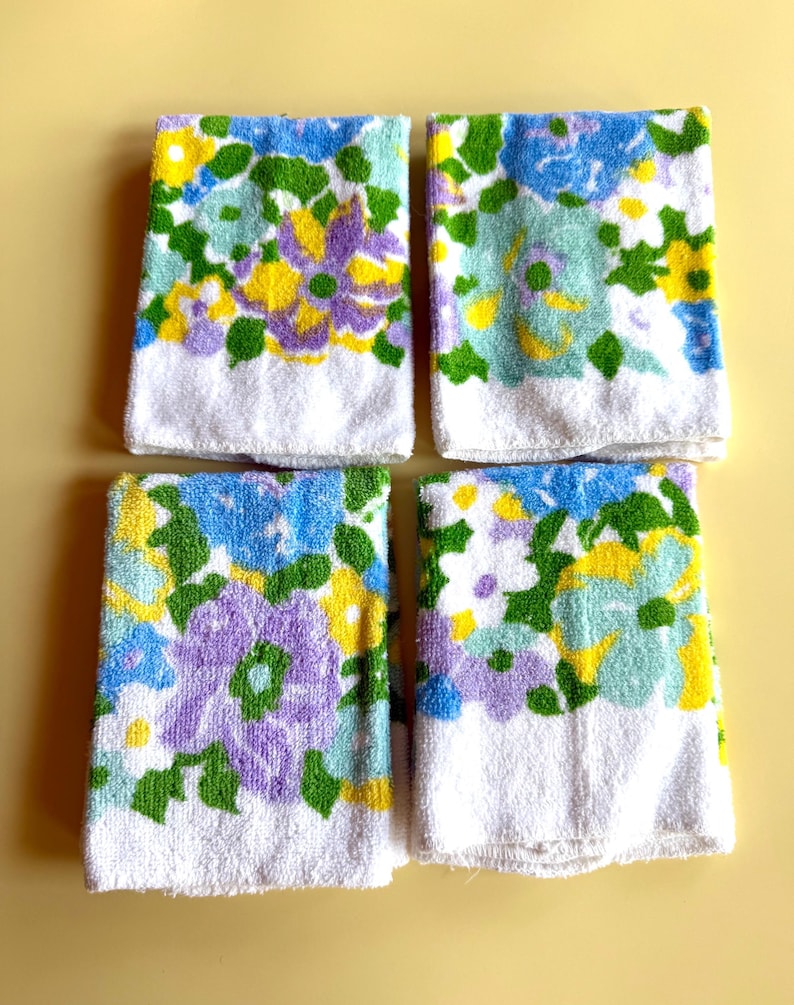 May include: Four white hand towels with a floral pattern in shades of blue, purple, yellow, and green. The towels have a white border at the bottom. The towels are arranged on a yellow background.