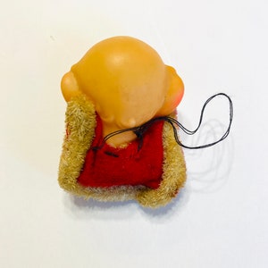 May include: A small, red and brown plush toy with a bald head and a string attached. The toy is sitting on a white surface.