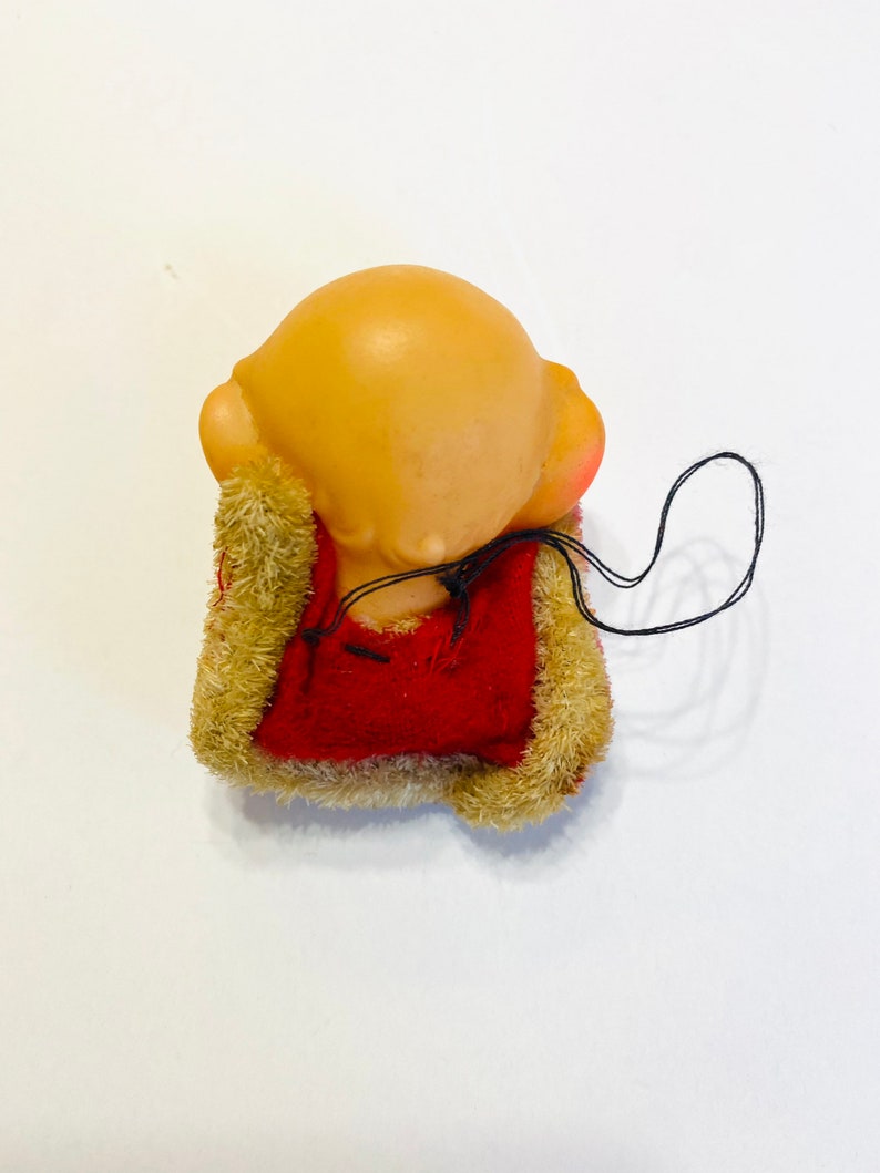 May include: A small, red and brown plush toy with a bald head and a string attached. The toy is sitting on a white surface.