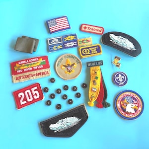May include: A collection of Boy Scout patches and pins on a blue background. Patches include an American flag, the words "Trained", "Webelos", and "205". Other patches feature eagles, alligators, and the Boy Scouts of America logo.