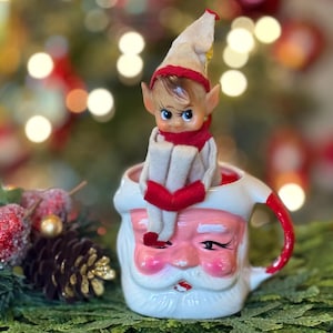 May include: A vintage Christmas elf doll with a red scarf and hat sits inside a Santa Claus mug. The mug is white with a pink Santa face and a red handle. The background is blurred with Christmas lights.