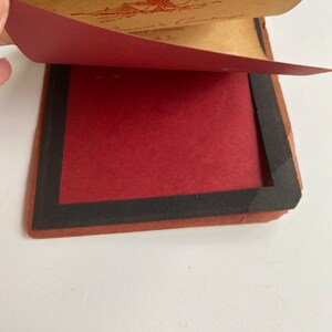 May include: A vintage greeting card with a red and brown color scheme. The card is open, revealing a red interior and a brown exterior with a red illustration and text. The card has a black border.
